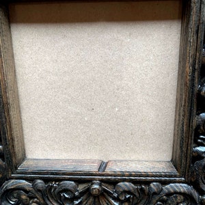May include: A dark brown wooden picture frame with ornate carvings around the edges. The frame has a rectangular opening with a light brown cardboard backing.
