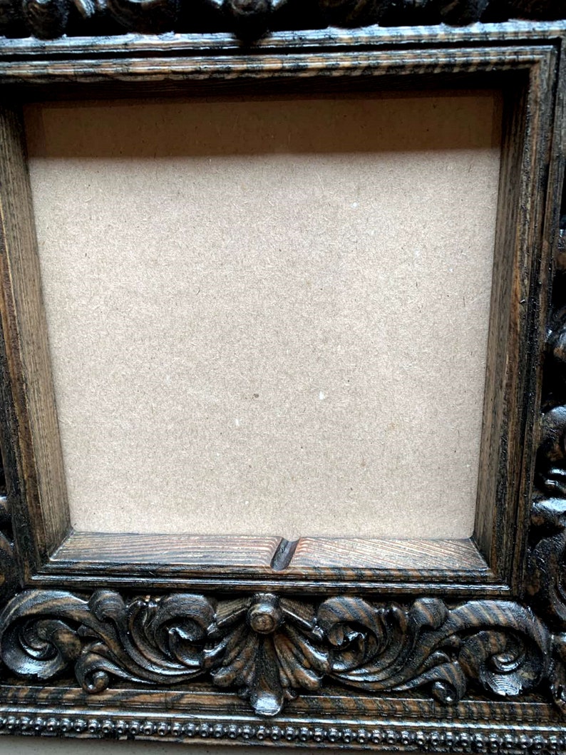 May include: A dark brown wooden picture frame with ornate carvings around the edges. The frame has a rectangular opening with a light brown cardboard backing.
