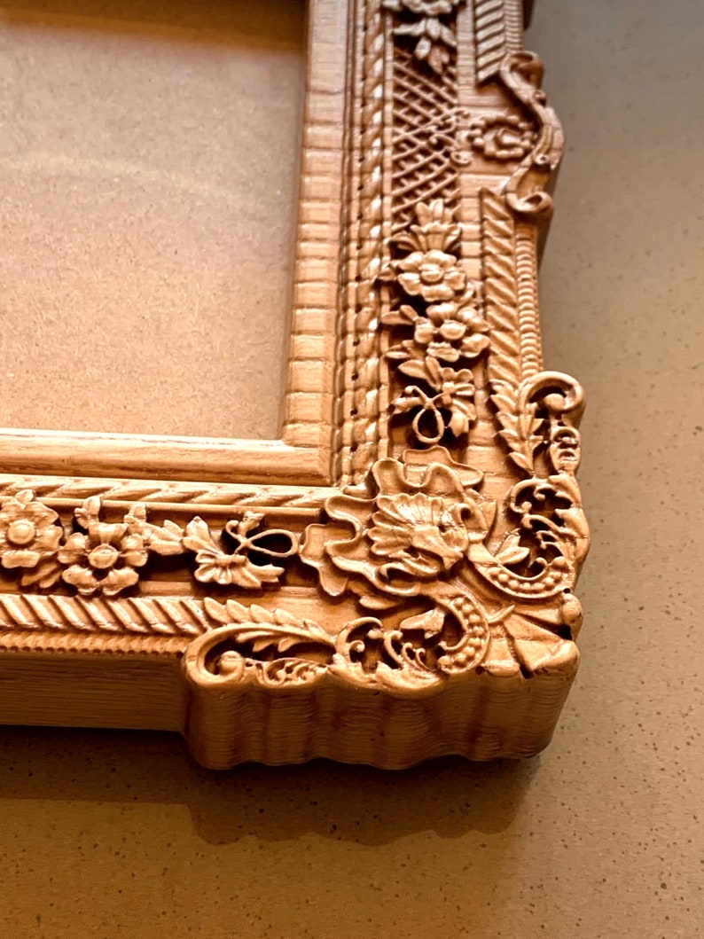 May include: A wooden picture frame with ornate carvings and a light brown finish. The frame has a raised edge and intricate floral designs.