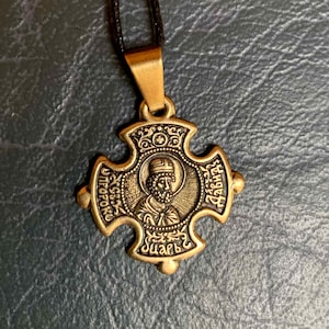 Puede incluir: Colgante en forma de cruz de color bronce, con la imagen de un hombre barbudo en un marco circular. El colgante cuelga de un cordón negro. La cruz presenta detalles ornamentados y texto.