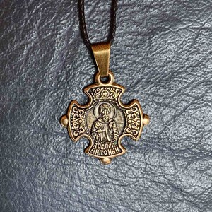 May include: A gold-toned cross pendant necklace with a black cord. The cross features a detailed design with a small image of a saint inside. The inscription on the cross reads "Св. Прп. Антоний".