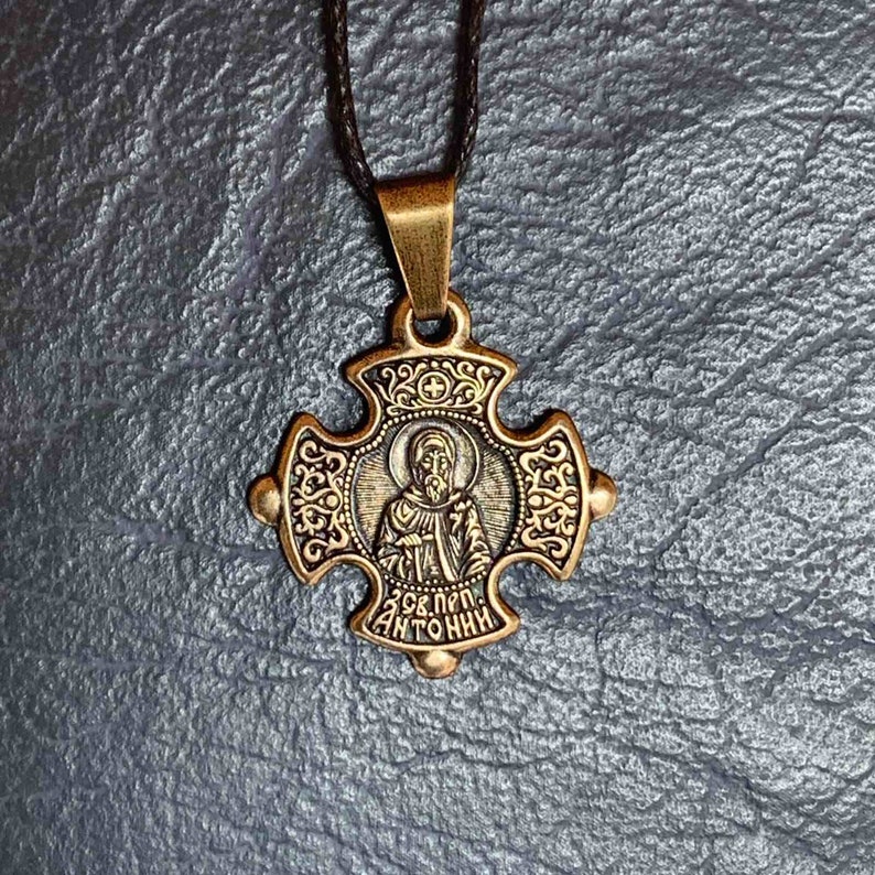 May include: A gold-toned cross pendant necklace with a black cord. The cross features a detailed design with a small image of a saint inside. The inscription on the cross reads "Св. Прп. Антоний".