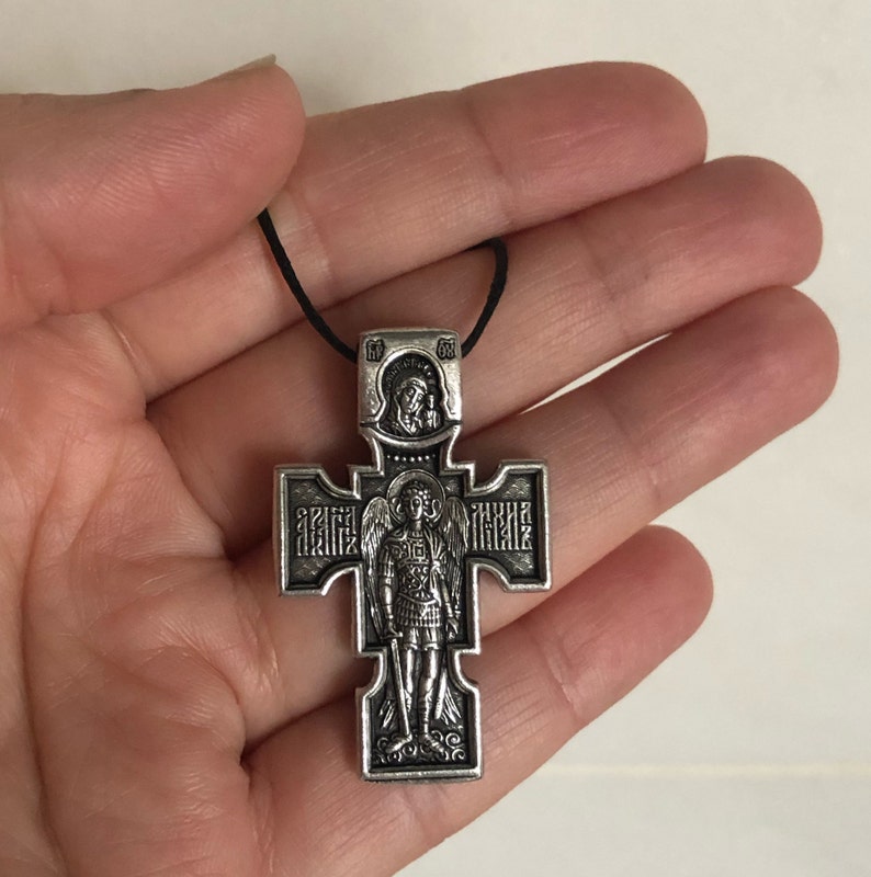 K&ouml;nnte beinhalten: Ein silberfarbener Anh&auml;nger in Form eines Kreuzes mit einem Bild eines Engels in der Mitte. Das Kreuz hat eine schwarze Schnur daran befestigt.