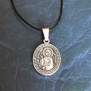 Op de afbeelding: Zilveren ovale hangerketting met een zwart koord. De hanger heeft een religieus icoon van een vrouw met een halo en de tekst "СВ. ЕКАТ. КСЕН. ПЕТЕРБУРГСКАЯ" rond de rand.