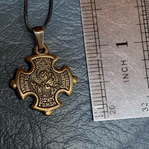 May include: A gold-toned metal pendant with a cross design. The pendant features a depiction of a saint with a crown and the text "St. Gregory the Great" and "Roman" in a circular frame. The pendant is approximately 1 inch in diameter.