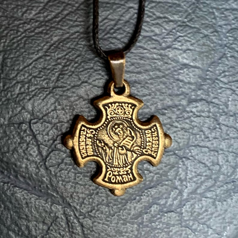 May include: A gold-toned metal pendant in the shape of a cross with a depiction of a religious figure inside. The cross has the words "Св. АФЕНО" and "Св. ГЕОРГИЙ" on the top and bottom arms, and "РОМАН" on the bottom. The pendant is hanging from a black cord.