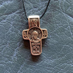 Könnte beinhalten: Ein bronzefarbener Anhänger in Form eines Kreuzes mit religiöser Ikonografie und kyrillischem Text. Das Kreuz hängt an einer schwarzen Kordel. Das Design umfasst eine zentrale Figur und aufwendige Details.