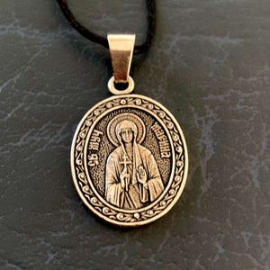 Può includere: Collana con ciondolo in argento con un'immagine rotonda di una donna che tiene una croce. L'immagine è circondata da un bordo decorativo con il testo "Св. Марина" in cirillico.