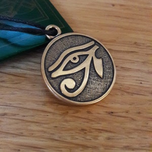 Puede incluir: Un colgante de color dorado con una imagen detallada del Ojo de Horus, un antiguo símbolo egipcio de protección y buena salud.
