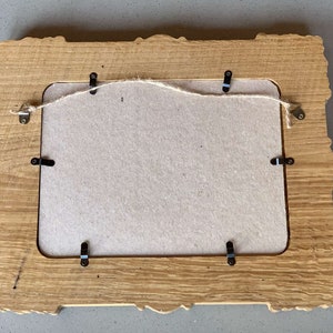 May include: A wooden picture frame with a carved design and a light brown finish. The frame has a cardboard backing and metal hanging hardware.