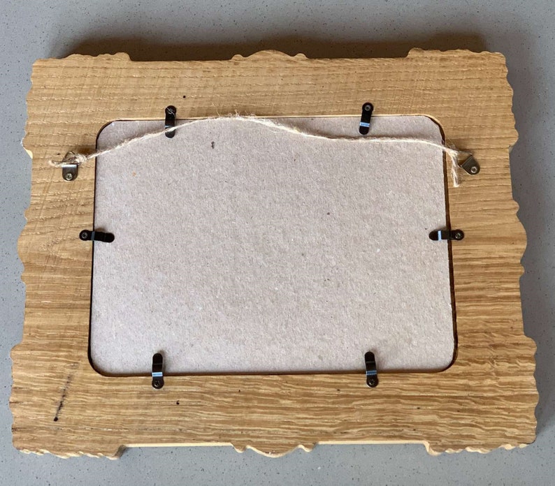 May include: A wooden picture frame with a carved design and a light brown finish. The frame has a cardboard backing and metal hanging hardware.
