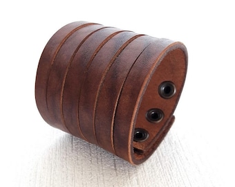 May include: A wide brown leather cuff bracelet with multiple horizontal bands. The bracelet has a distressed appearance and features three black metal snap closures. The leather is a rich, warm brown color.