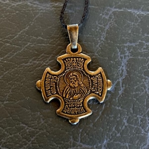 Może przedstawiać: Złoty metalowy wisiorek w kształcie krzyża z wizerunkiem brodatego mężczyzny w środku. Wisiorek ma wygrawerowane słowa "Св. Сергий" i "Радонежский".