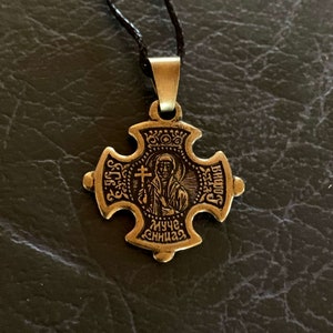 May include: A gold-toned metal pendant in the shape of a cross with a small image of a saint in the center. The pendant has the words "муче. сница" inscribed on the bottom and "святой" inscribed on the right side.