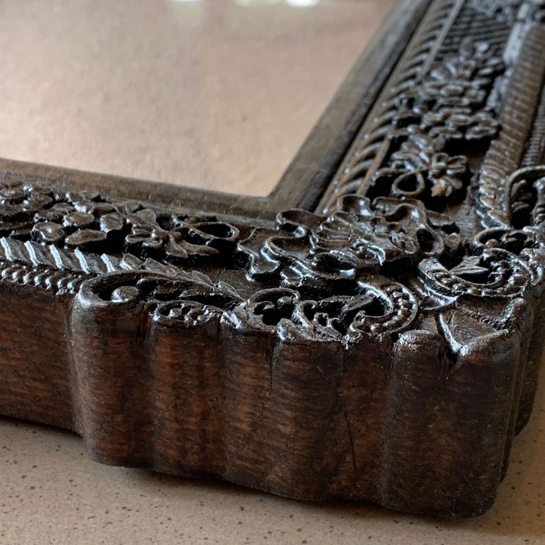 May include: Close-up of a dark brown wooden picture frame with intricate carved details. The frame has a scalloped edge and a floral design.