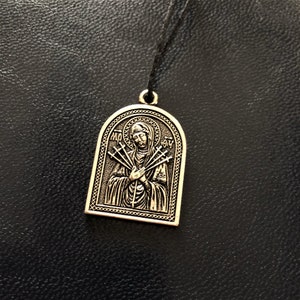 Peut inclure: Un pendentif en argent avec une image religieuse d'une femme avec sept épées qui lui percent le cœur. Le pendentif est sur un cordon noir.