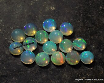 May include: A group of 14 round, iridescent opal cabochons in various shades of green, blue, and yellow. The cabochons are arranged in a loose cluster on a dark gray surface. Gemstonedealers is printed in white on the surface.