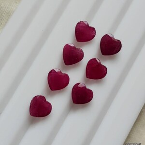 May include: Six small, faceted heart-shaped gemstones in a deep, vibrant magenta-red color. The stones are arranged in a staggered, diagonal pattern, showcasing their polished surfaces and distinct heart silhouettes against a white background.