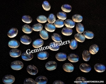 May include: A collection of oval-shaped moonstone cabochons, with a milky white color and iridescent blue flashes. The stones are scattered on a black background.