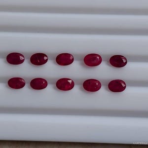 May include: Ten oval-cut, deep red rubies arranged in two horizontal rows of five. Each gemstone features a faceted surface that reflects light, showcasing a consistent crimson hue and a polished finish, presented on a white grooved display tray.