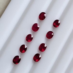 Può includere: Dieci rubini rosso intenso dal taglio ovale disposti in due file verticali parallele. Ogni gemma presenta una superficie sfaccettata che riflette la luce, mostrando una vibrante tonalità cremisi e una finitura lucida su una superficie bianca.