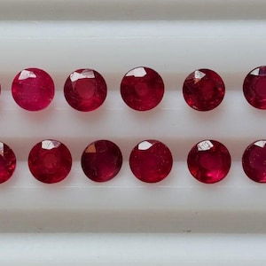 May include: A collection of eighteen round, faceted rubies arranged in two rows. The gemstones are a deep, rich red color, with visible facets catching the light. The rubies are displayed on a white surface.
