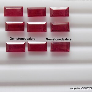 May include: Nine rectangular red gemstones arranged in three rows of three. The gemstones are on a white surface with a striped pattern. The text "Gemstonedealers" appears twice on the surface.