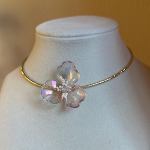 May include: A delicate necklace featuring a three-petal flower pendant. The pendant has an iridescent, translucent appearance with white accents. The necklace has a thin, gold-colored chain, displayed on a white mannequin.