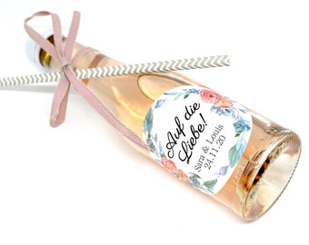 May include: A small bottle of pink sparkling wine with a floral patterned label that says "Auf die Liebe!"  Sara & Louis 24.11.20. The bottle is tied with a pink ribbon and a striped straw.