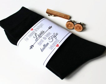 May include: A white gift tag with the text "For meinen Bräutigam Louis damit du keine kalten Füße bekommst" with a red heart and arrow design. The tag is wrapped around a pair of black socks. The image also shows a wooden tie clip and cufflinks with a camera design.