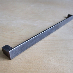 May include: A long, rectangular, metal drawer pull with a brushed finish. The pull is made of a dark gray metal and has a slightly textured surface.