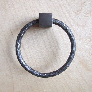 Peut inclure: Une poignée de porte en métal martelé en forme d'anneau avec une base carrée. La poignée est de couleur gris foncé.