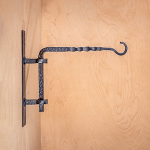 May include: Black metal wall-mounted hook with a decorative scroll design. The hook is attached to a wooden wall.