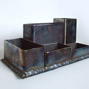 Puede incluir: Un organizador de escritorio de metal rústico con cinco compartimentos. El organizador está hecho de metal oscuro con un acabado desgastado.