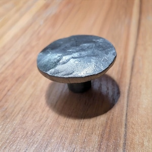 May include: A round, silver-colored metal knob with a textured surface. The knob is mounted on a wooden surface.