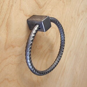 May include: A metal cabinet pull with a square base and a circular handle. The handle is made of twisted metal, and the pull is mounted on a light wood surface. The hardware has a rustic, industrial aesthetic.