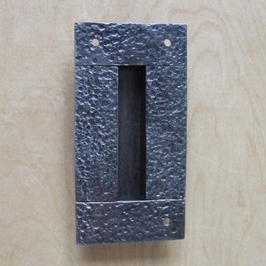 May include: A silver-toned metal door pull with a textured finish. The pull has a rectangular shape with a vertical slot in the center.