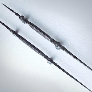 Może przedstawiać: Dwa czarne metalowe uchwyty do drzwi o skręconym wzorze. Uchwyty są długie i smukłe, ze spiczastym końcem.