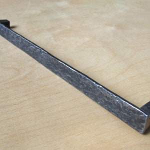 Puede incluir: Un tirador de cajón largo, rectangular, de metal martillado con extremos cuadrados. El tirador es de color gris oscuro.