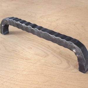 Könnte beinhalten: Ein schwarzer Metall-Schrankgriff mit einem strukturierten, wellenförmigen Design. Der Griff ist gebogen und hat einen einfachen, modernen Stil.