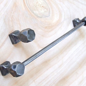 Peut inclure: Une barre de serviette en métal noir avec deux boutons décoratifs hexagonaux à facettes. La barre est montée sur une surface en bois clair.