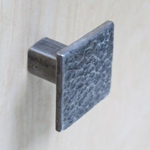 May include: A square, silver-toned cabinet knob with a textured surface.