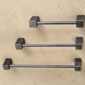 May include: Three metal towel bars with square ends. The bars are different lengths and are arranged in a descending order from top to bottom.
