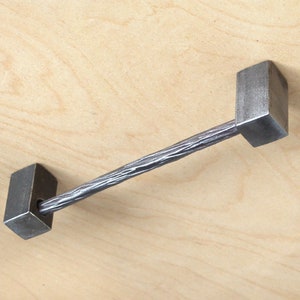 May include: A silver metal drawer pull with a textured bar and square ends. The pull is mounted on a light wood surface.