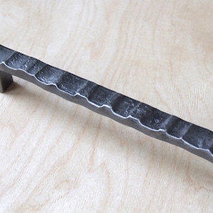 May include: A black metal cabinet pull with a textured, hammered finish. The pull has a rectangular shape with rounded edges.
