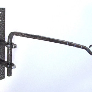 May include: A hand-forged, dark metal wall-mounted plant hanger. The hanger features a textured, rectangular mounting plate with two square brackets and a curved arm ending in a decorative hook. The metal has a hammered finish.