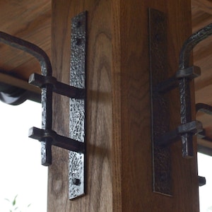 Może przedstawiać: Dwa czarne metalowe wsporniki są zamontowane na drewnianym słupie. Każdy wspornik ma ozdobny wzór i jest przymocowany do metalowego pręta.