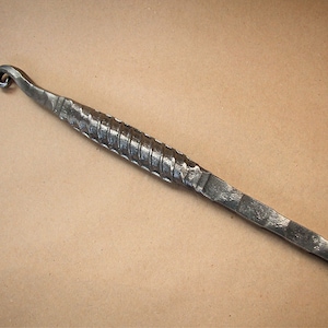May include: A long, thin, metal tool with a twisted handle and a hook at one end. The tool is made of dark metal and has a rustic appearance.