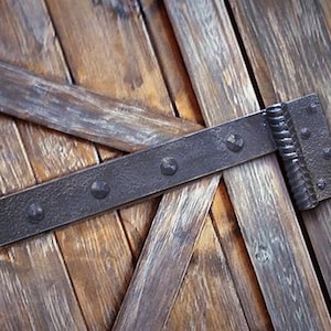 Puede incluir: Primer plano de una bisagra de metal negro con detalles decorativos fijada a una puerta de madera desgastada.