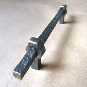 May include: A black metal cabinet pull with a square shape and a knotted design. The pull is made of a textured metal and has a rustic look.